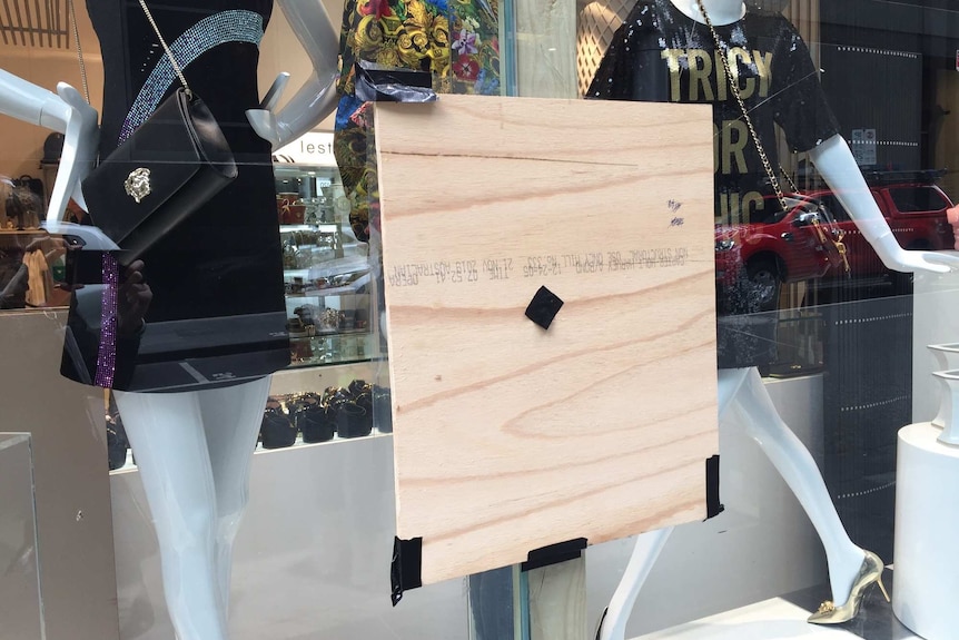 A shop window with dressed mannequins on show has a small wooden square attached to the glass to cover a hole.