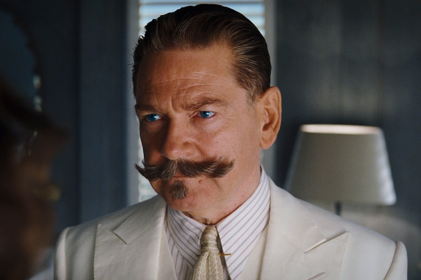 A 60-something man with a twirly moustache and slicked-back hair, wearing a linen suit, looks stern and determined