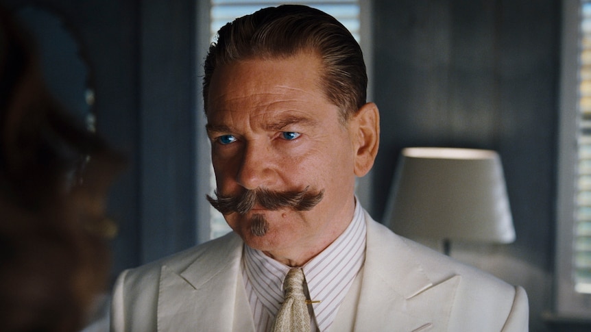 A 60-something man with a twirly moustache and slicked-back hair, wearing a linen suit, looks stern and determined