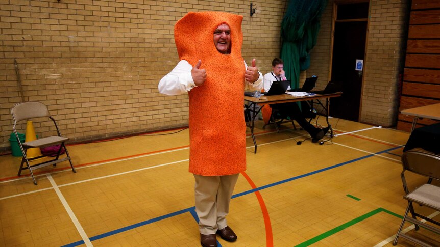 A man in a a bright orange fishfinger suit gestyres with both thumbs up at a polling booth in a basketball court.