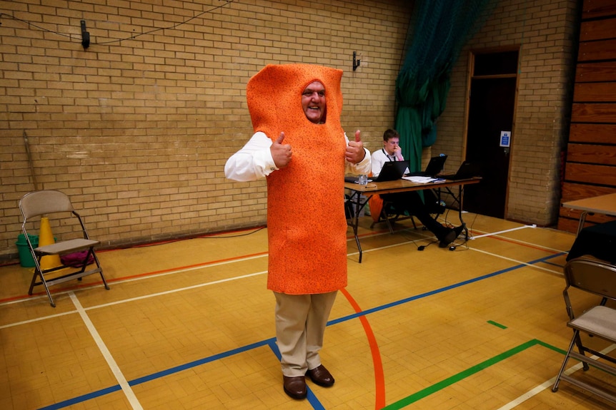 A man in a a bright orange fishfinger suit gestyres with both thumbs up at a polling booth in a basketball court.