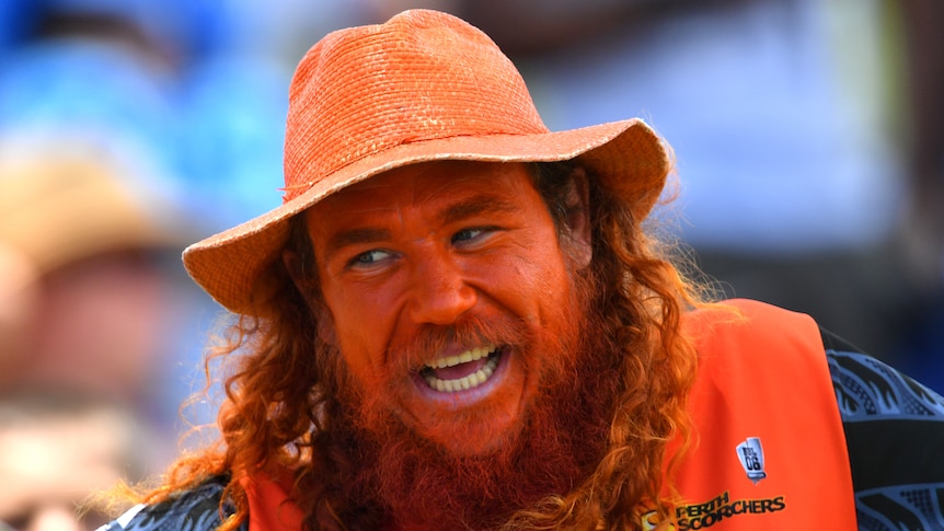 A man with long red hair wearing an orange singlet, hat and hair colour