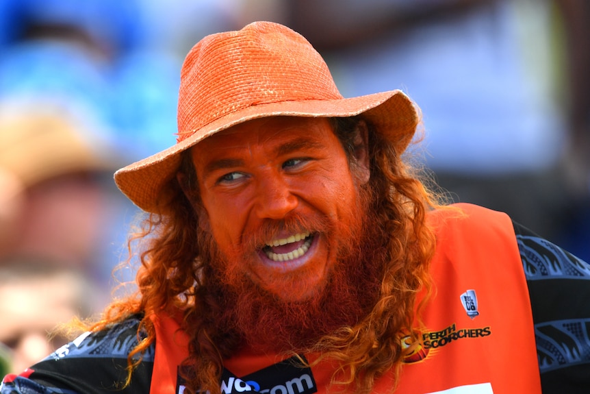 A man with long red hair wearing an orange singlet, hat and hair colour