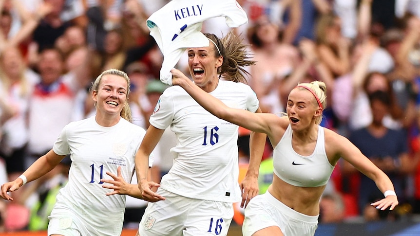 Three female soccer players are celebrating a win by running across a soccer field beaming. A crowd is behind them. 