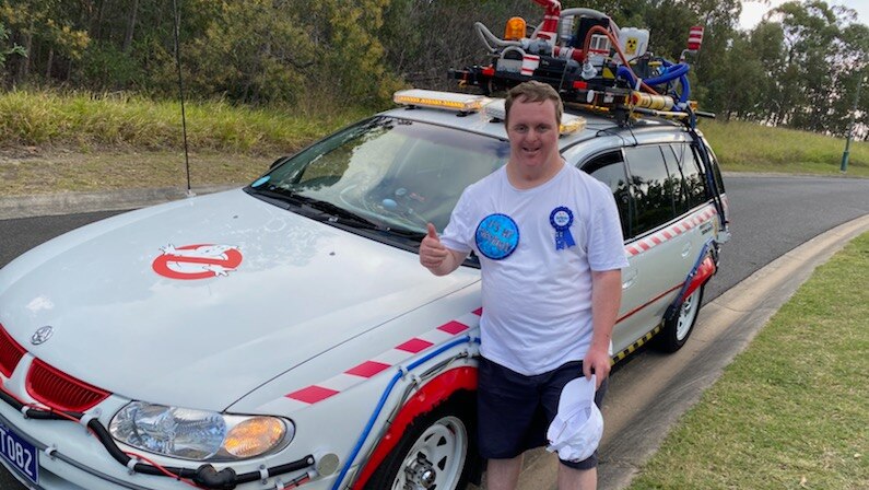 man in white shirt with birthday boy badge leans on a ghostbusters car with his thumbs up