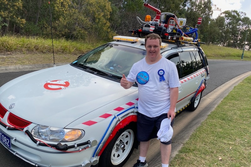 man in white shirt with birthday boy badge leans on a ghostbusters car with his thumbs up