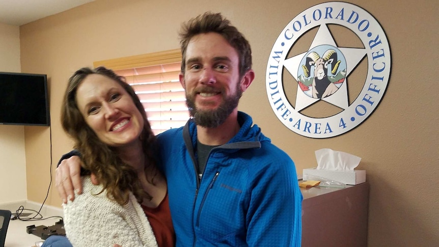 Two Caucasian people look into a camera in front of a large 'Colorado Wildlife Area 4 Office' logo—a star with a mountain goat.