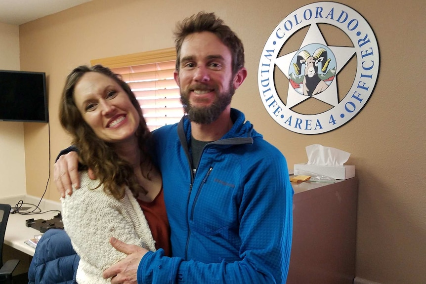 Two Caucasian people look into a camera in front of a large 'Colorado Wildlife Area 4 Office' logo—a star with a mountain goat.