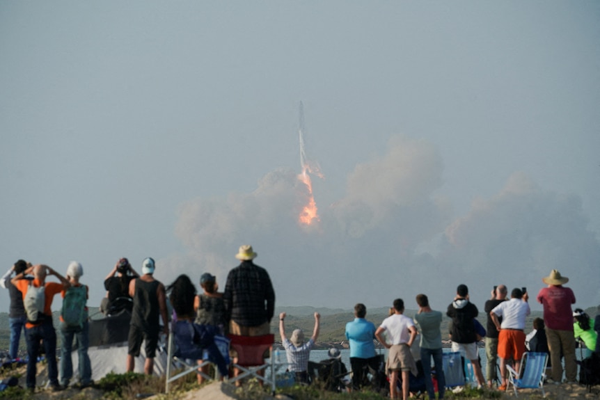 A crowd of people watching a rocket launch out of plumes of smoke