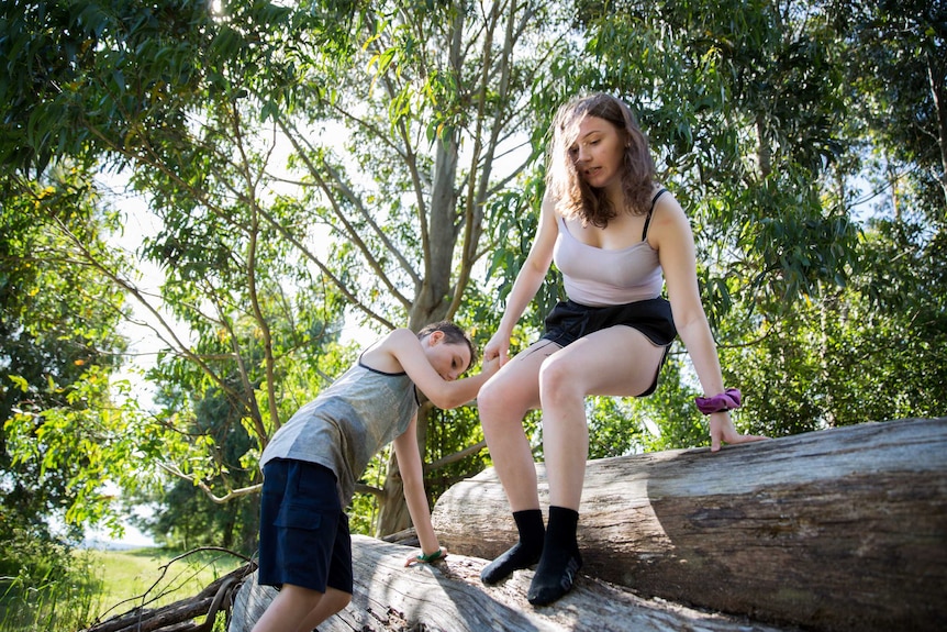 A girl holds onto a younger boy's hand as she climbs down a pile of old logs.