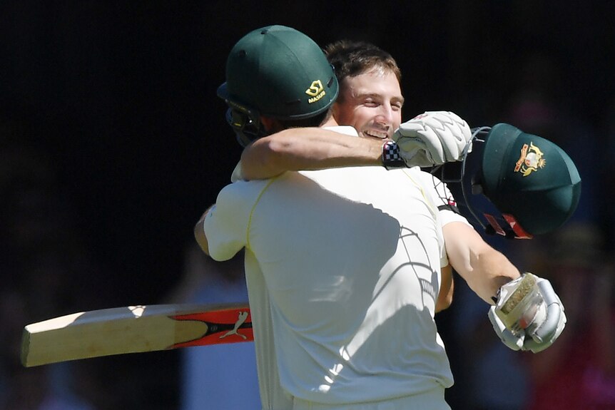Two cricket players embrace, one with his helmet off. 