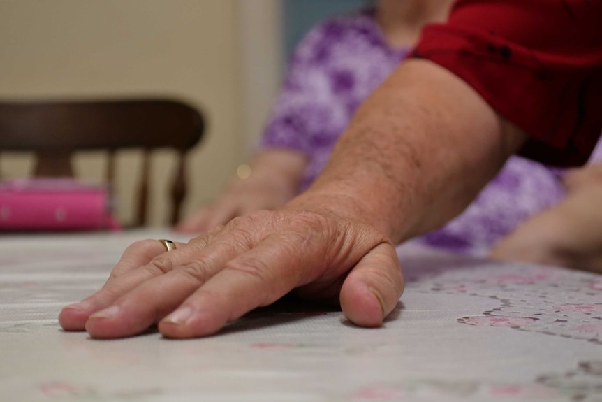 An aging man's hand is in focus, he wears a ring. Out of focus in the background is a woman in a purple shirt.
