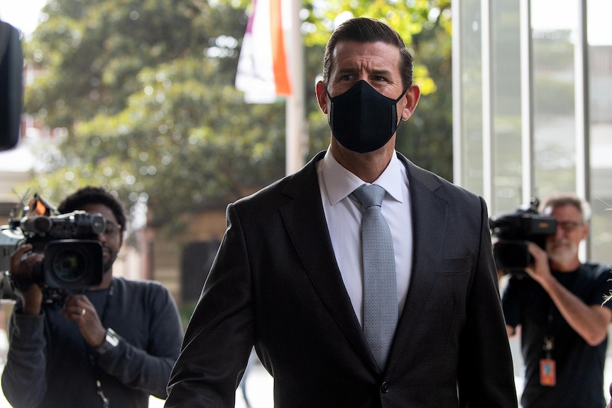 A man wearing a suit and a black face mask flanked by television cameras. 