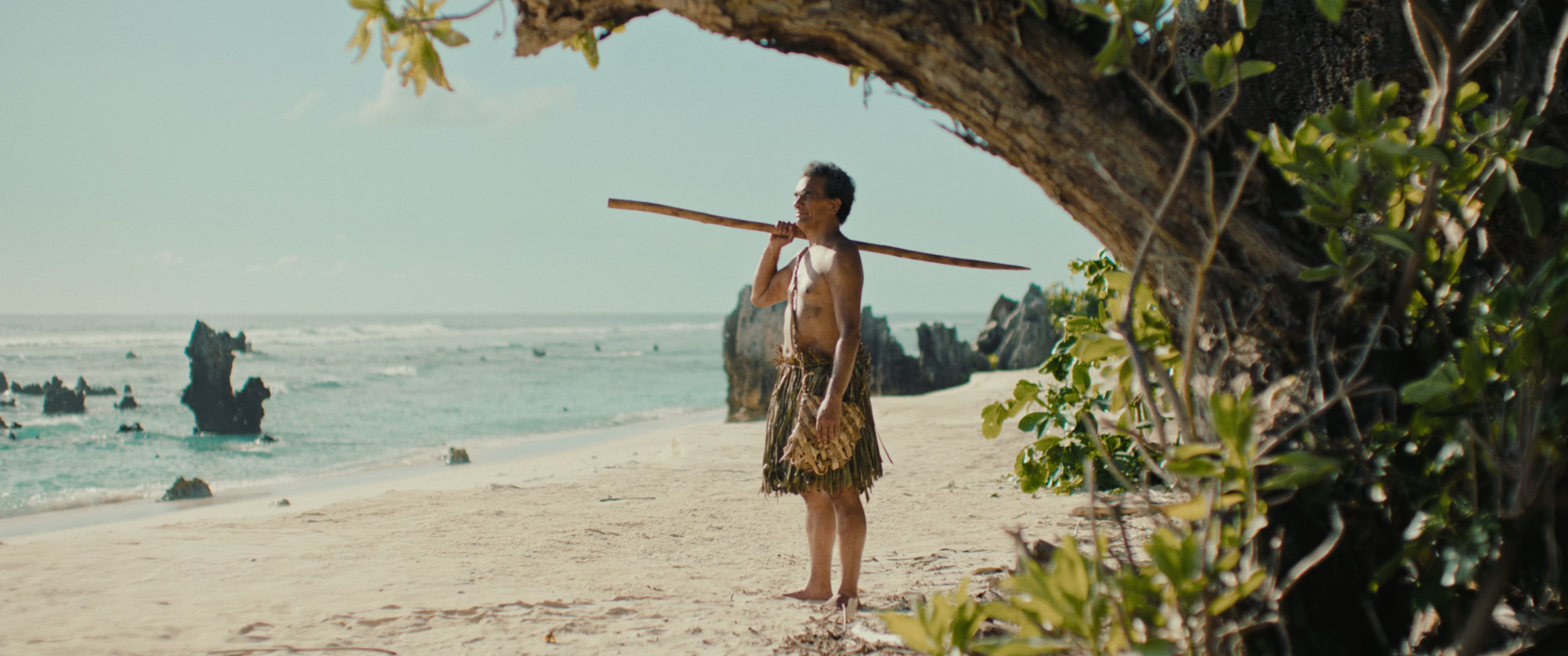 A man wearing traditional wear from Naoero stands a beach looking out to sea.