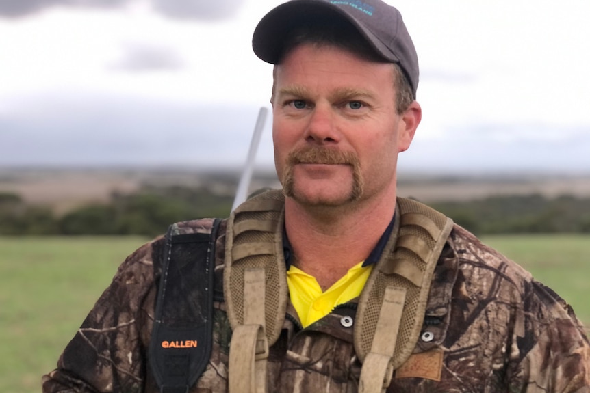 A man with a handlebar moustache wearing camouflage looks at the camera.