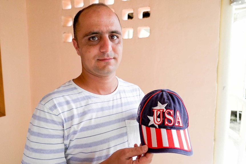 A male asylum seeker stands inside a concrete room holding a baseball cap with USA written on it.