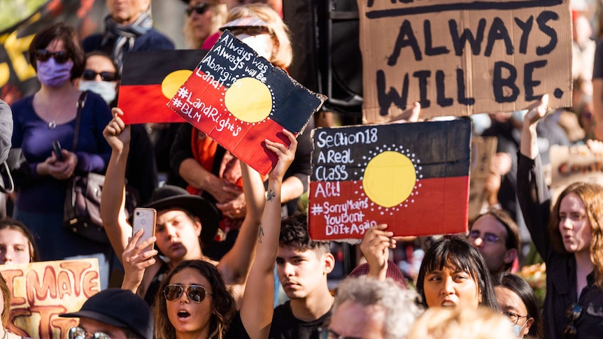 A group of protestors hold up signs saying 'always was always will be Aboriginal land', coloured in as Aboriginal flags.