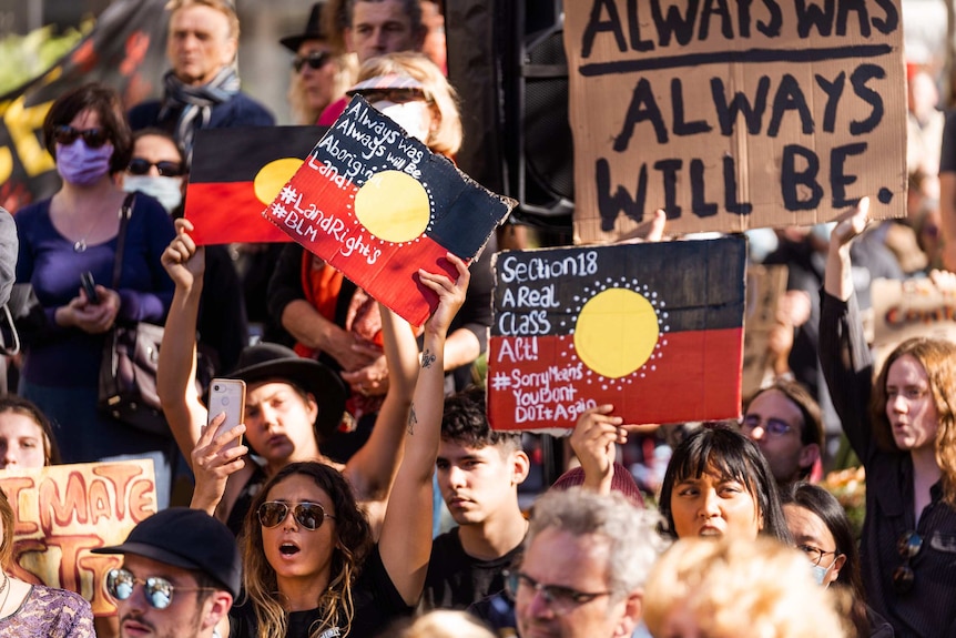 A group of protestors hold up signs saying 'always was always will be Aboriginal land', coloured in as Aboriginal flags.
