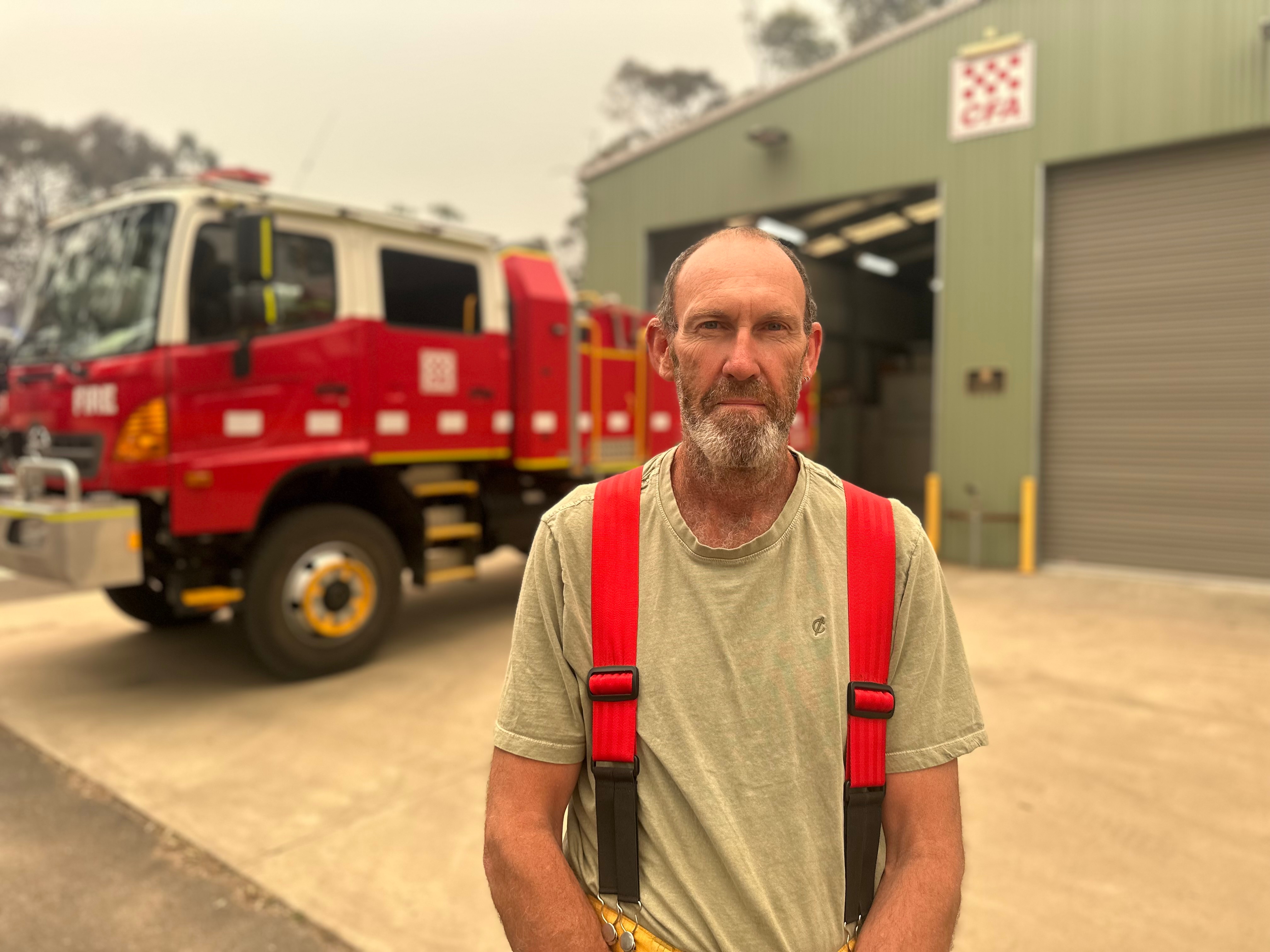 Steve Field has a beard and wears red braces over a tshirt and stands near a red fire truck and shed with a CFA sign.