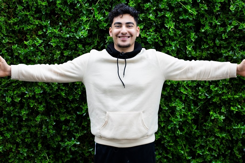 A young man in a hoodie holds his arms out in front of a hedge