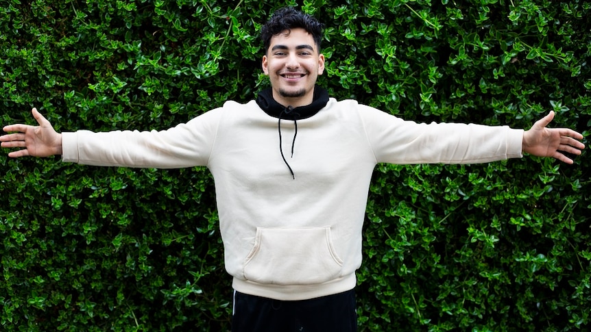 A young man in a hoodie holds his arms out in front of a hedge