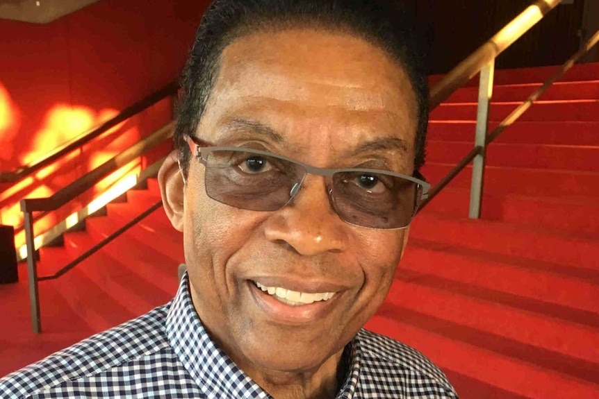 A man wearing a checked shirt and glasses stands at the foot of red-carpeted stairs.