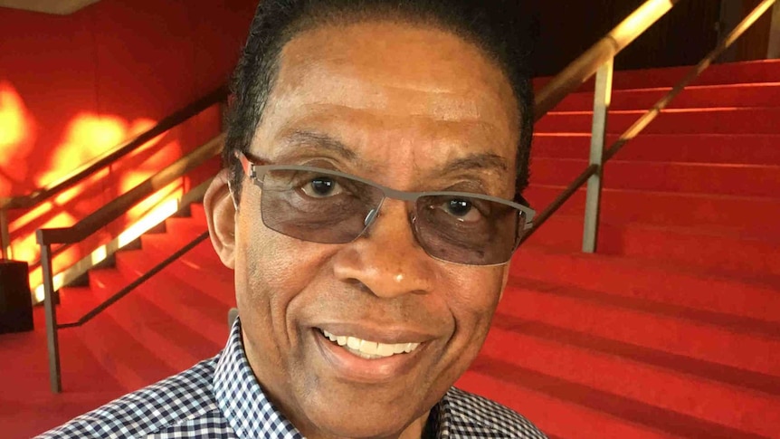 A man wearing a checked shirt and glasses stands at the foot of red-carpeted stairs.