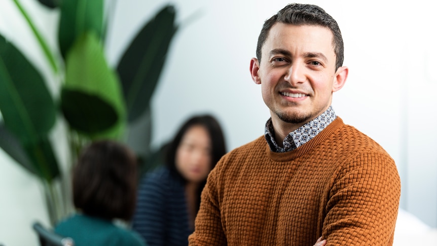 A man wearing a brown jumper standing in front of a plant. 