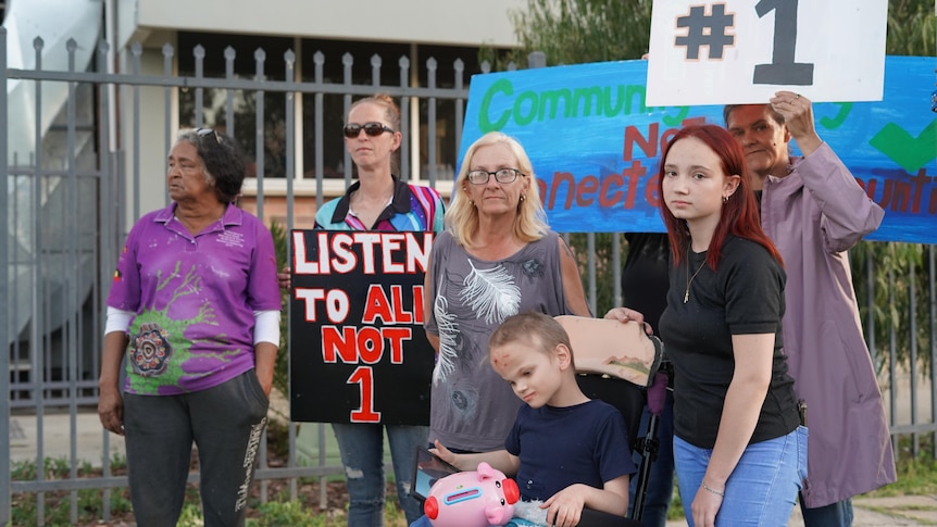 A group of women surround a boy in a wheelchair in front of a fenced building