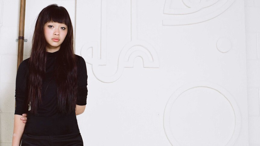 A portrait of Angie Pai wearing all black in a white gallery space as she stands in front of an artwork of hers.