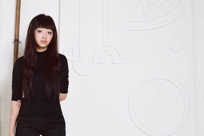 A portrait of Angie Pai wearing all black in a white gallery space as she stands in front of an artwork of hers.