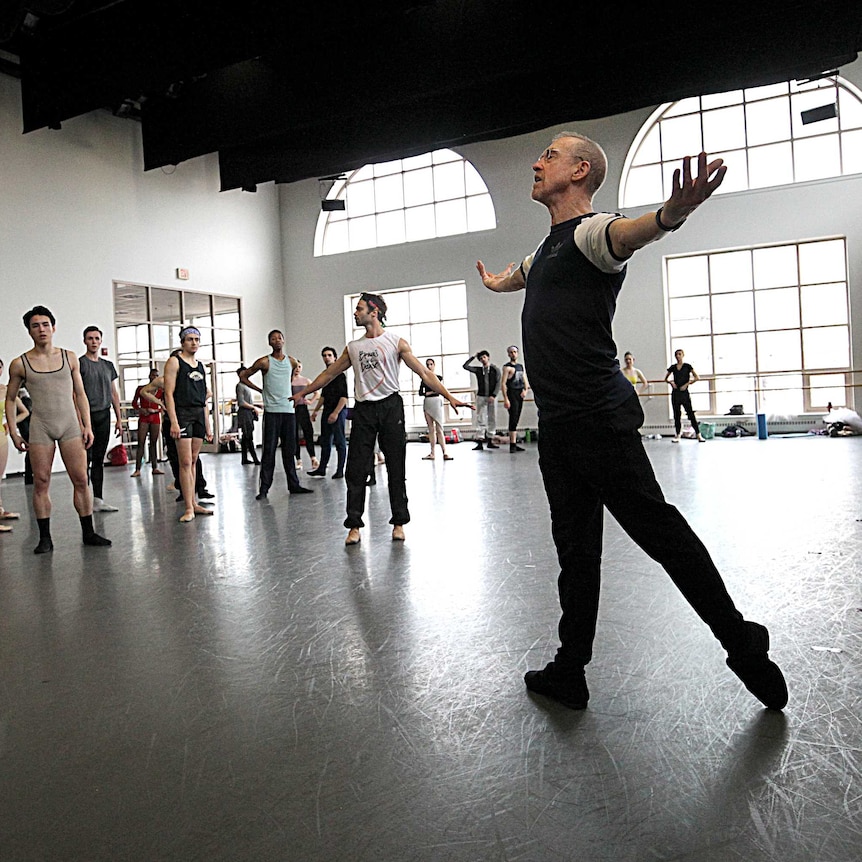 William Forsythe, with his arms outstretched, demonstrates a move to a group of dancers in a well-lit rehearsal room.