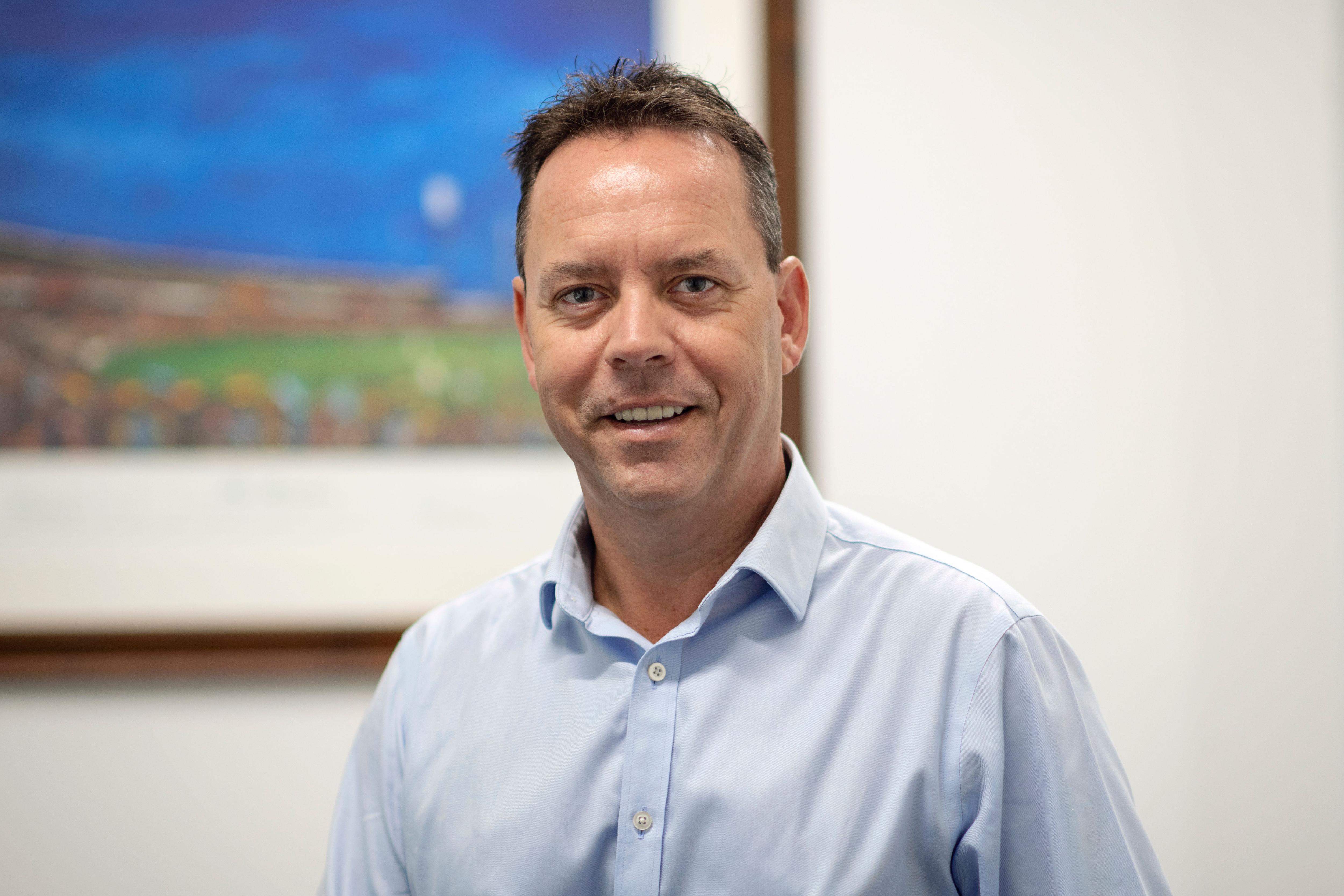 Corporate headshot of man smiling in a blue shirt in an office with a painting behind.