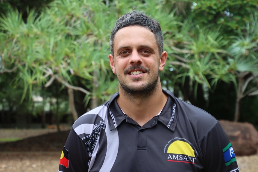 A man with short black hair aged in his 30s wearing a black collared shirt smiling at the camera