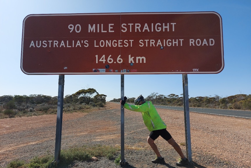 Man pushing one of the sign poles on a large "90 mile straight" brown tourism sign
