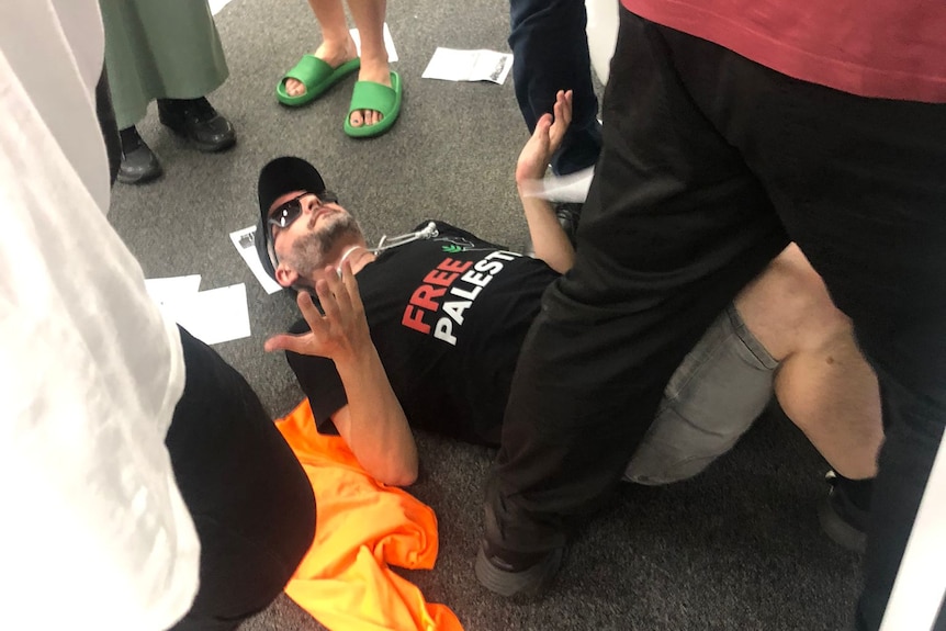A man in a dark cap lies on a floor, surrounded by people.