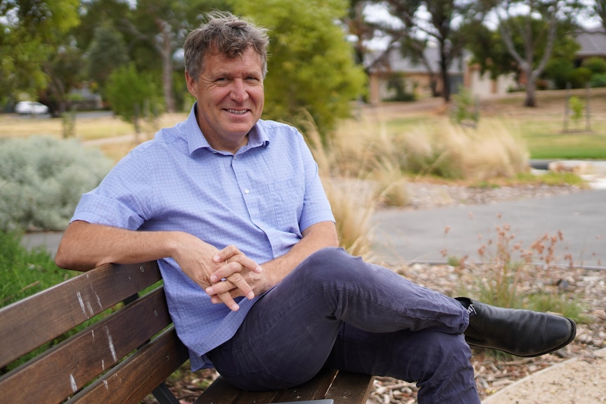 A man sits with his legs crossed and hands folded on a bench in a park