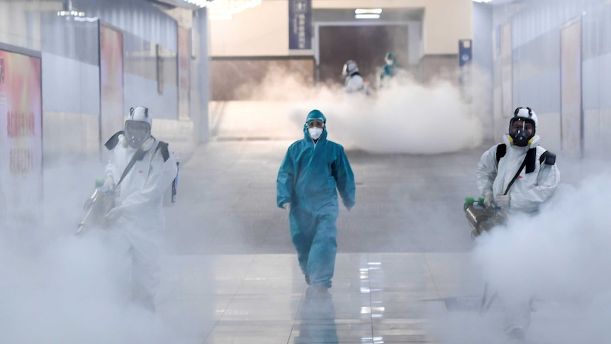 A person in a blue full body medical suit walks in a corridor flanked by two others spraying disinfectant in large white smoke.