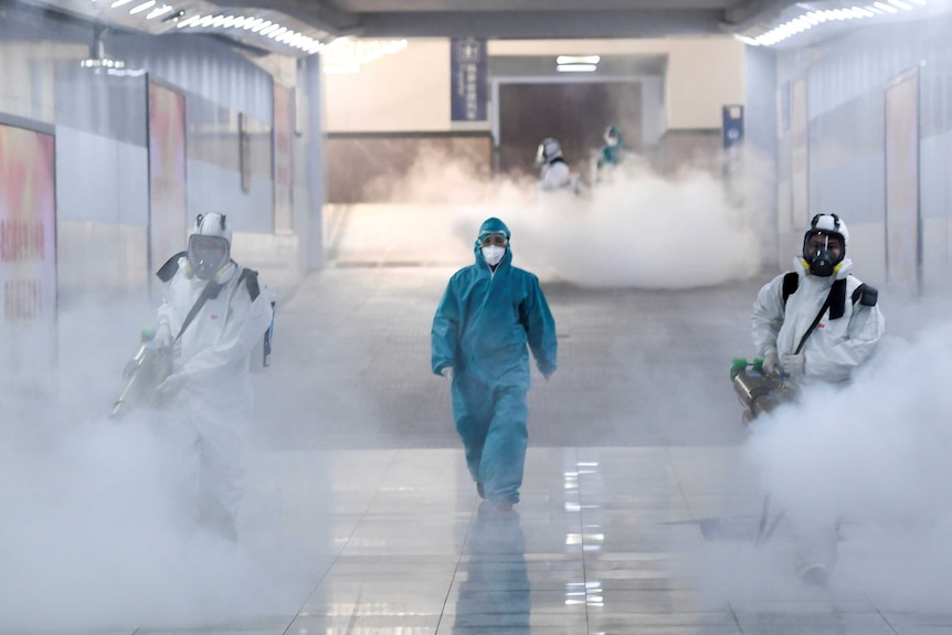 A person in a blue full body medical suit walks in a corridor flanked by two others spraying disinfectant in large white smoke.