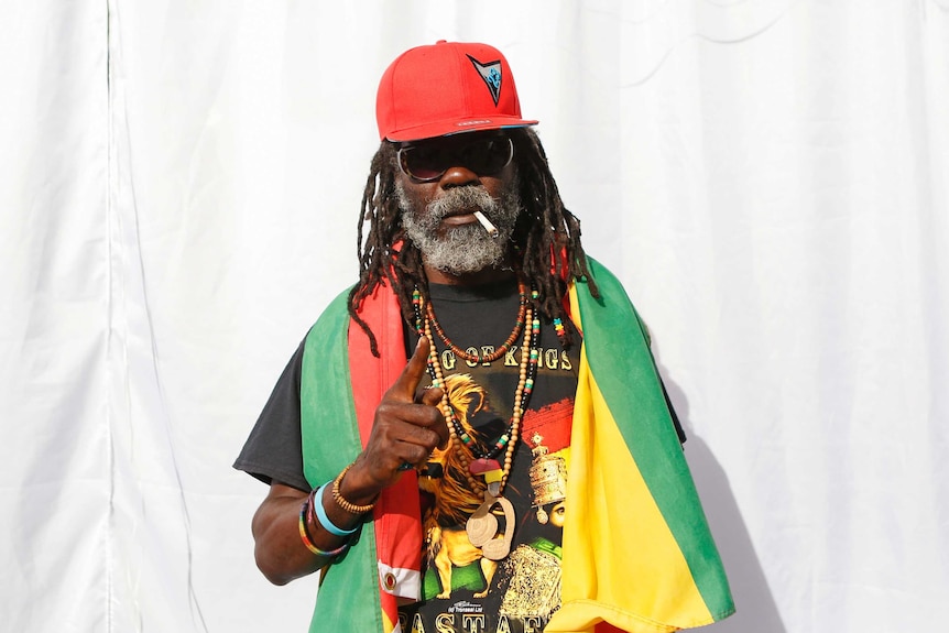 An elderly Rastafarian stands with a multicoloured flag around his neck.
