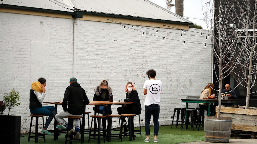 People sit outside at a Geelong venue, many wearing facemasks. There is a white wall behind them and a string of lights.