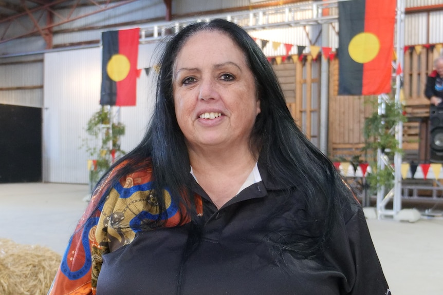 A woman wearing a block polo with long hair. Two Aboriginal flags on a stage with gum leaves in the background. 