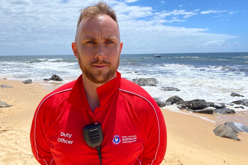 A man wearing  a red surf life saving jumper, standing in front of the beach. 