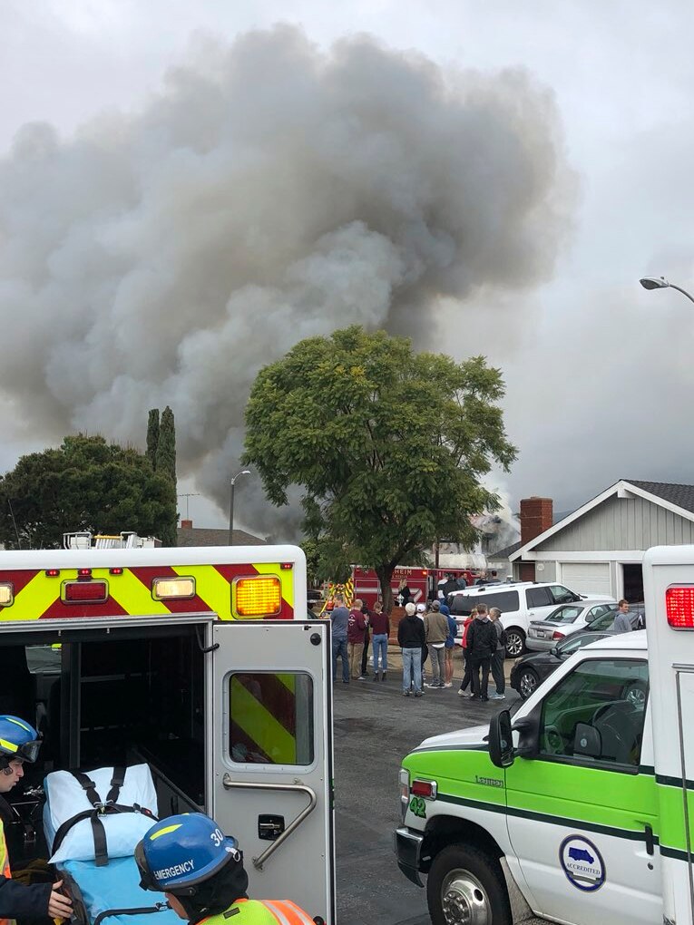 Smoke billows from a house in the distance as a crowd stands in front of two parked ambulances.