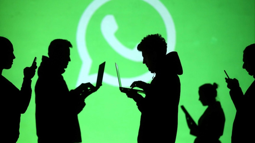 Silhouettes of laptop and mobile device users are seen next to a screen projection of Whatsapp logo