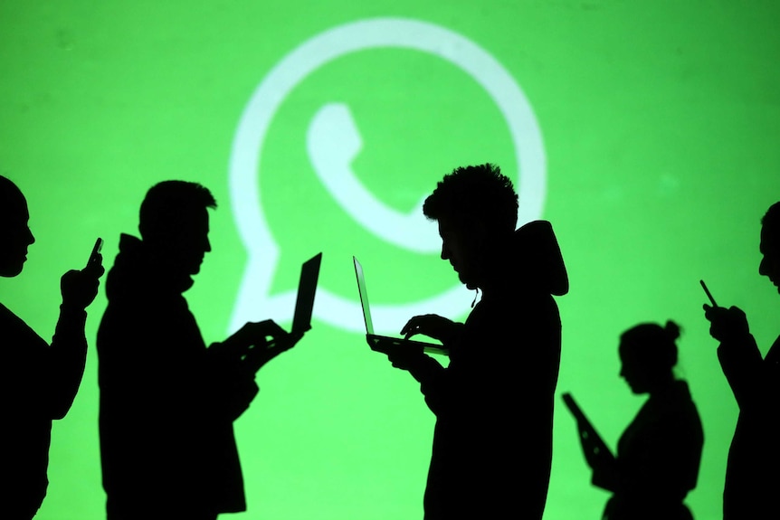 Silhouettes of laptop and mobile device users are seen next to a screen projection of WhatsApp logo