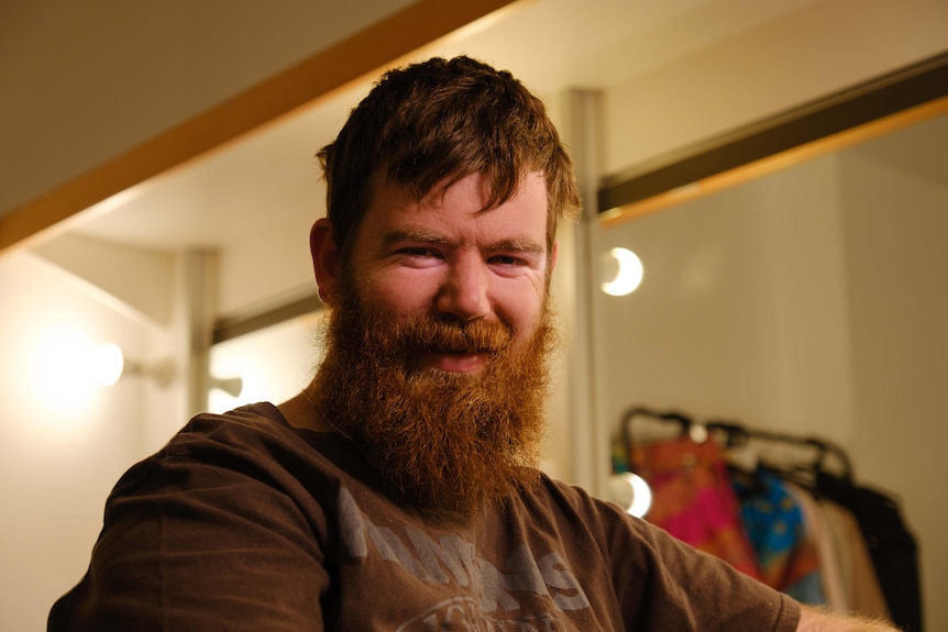 Scott Price with a long beard sits in a dressing room smiling at the camera.
