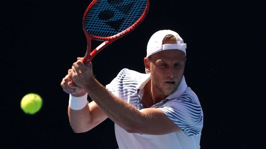A man in a white shirt and hat plays a backhand tennis shot.