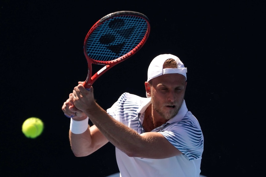 A man in a white shirt and hat plays a backhand tennis shot.