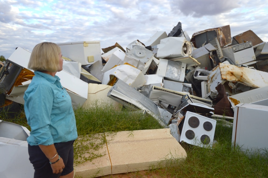 A lady with blonde hair in a blue shirt standing and looking to a large pile of white goods at a dump